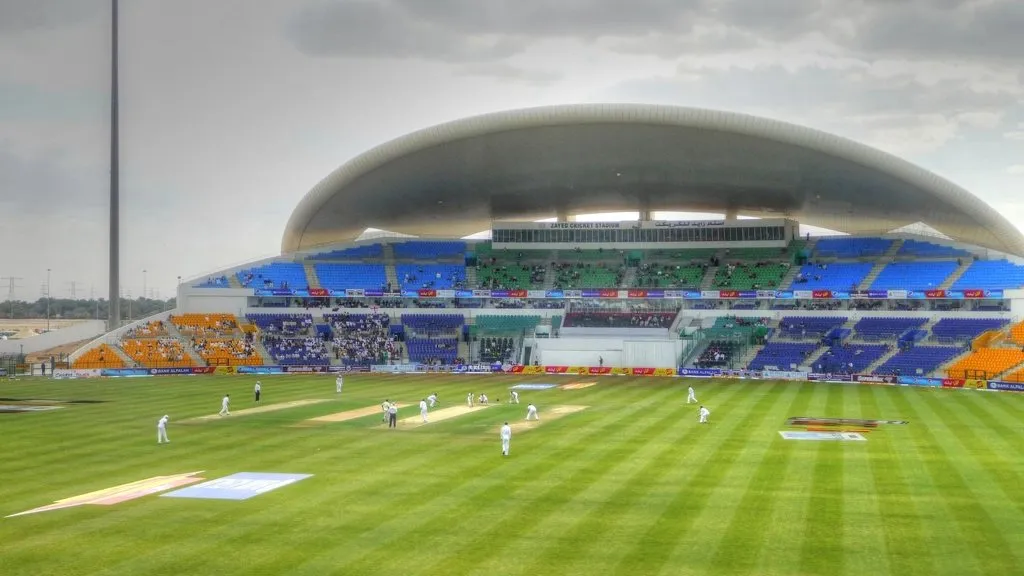 A cricket stadium during a match, showing the pitch square in the middle of the outfield